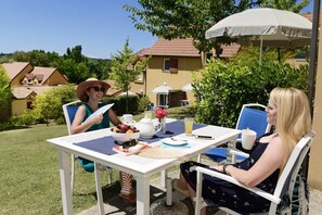 Un espace extérieur aménagé pour prendre les repas, avec une table dressée pour deux personnes, entouré de verdure et avec une maison en arrière-plan.