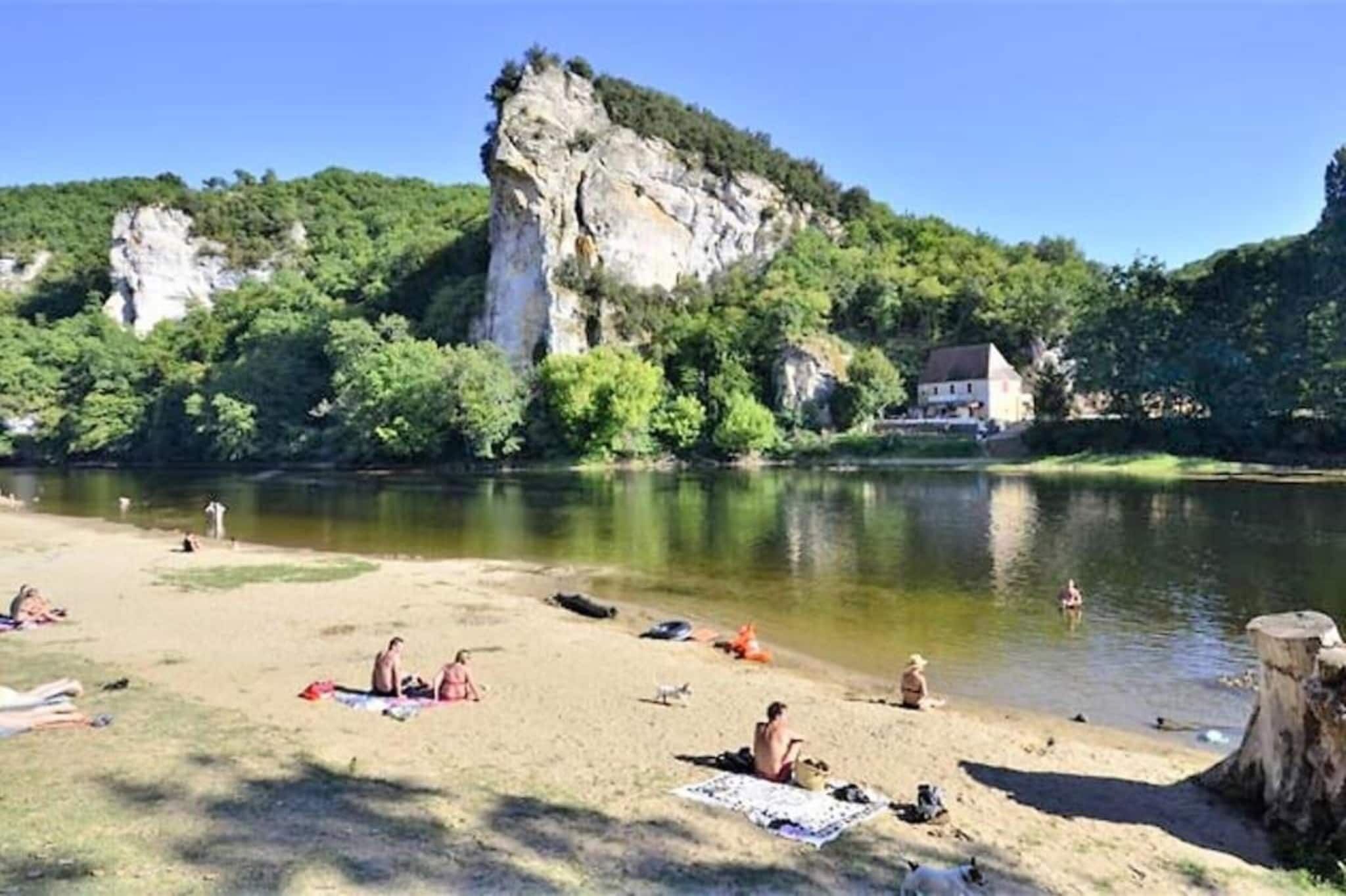Une plage au bord de la rivière où les gens se prélassent au soleil et se baignent, entourée de verdure et bordée d’une falaise imposante.