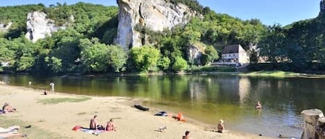 A riverside beach with people sunbathing and swimming, surrounded by greenery and a prominent cliff.