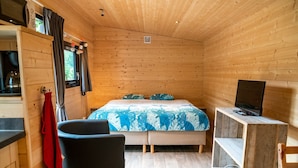 A wooden cabin room with a bed, a TV on a wooden stand, a red towel hanging, and a window with curtains. - Woodland Lodge Retreat in the Ardennes (Olloy-sur-Viroin)