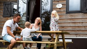 A family of four seated at an outdoor wooden picnic table in front of a wooden cabin with a sign that reads "Wool Lodge." - Woodland Lodge Retreat in the Ardennes (Olloy-sur-Viroin)