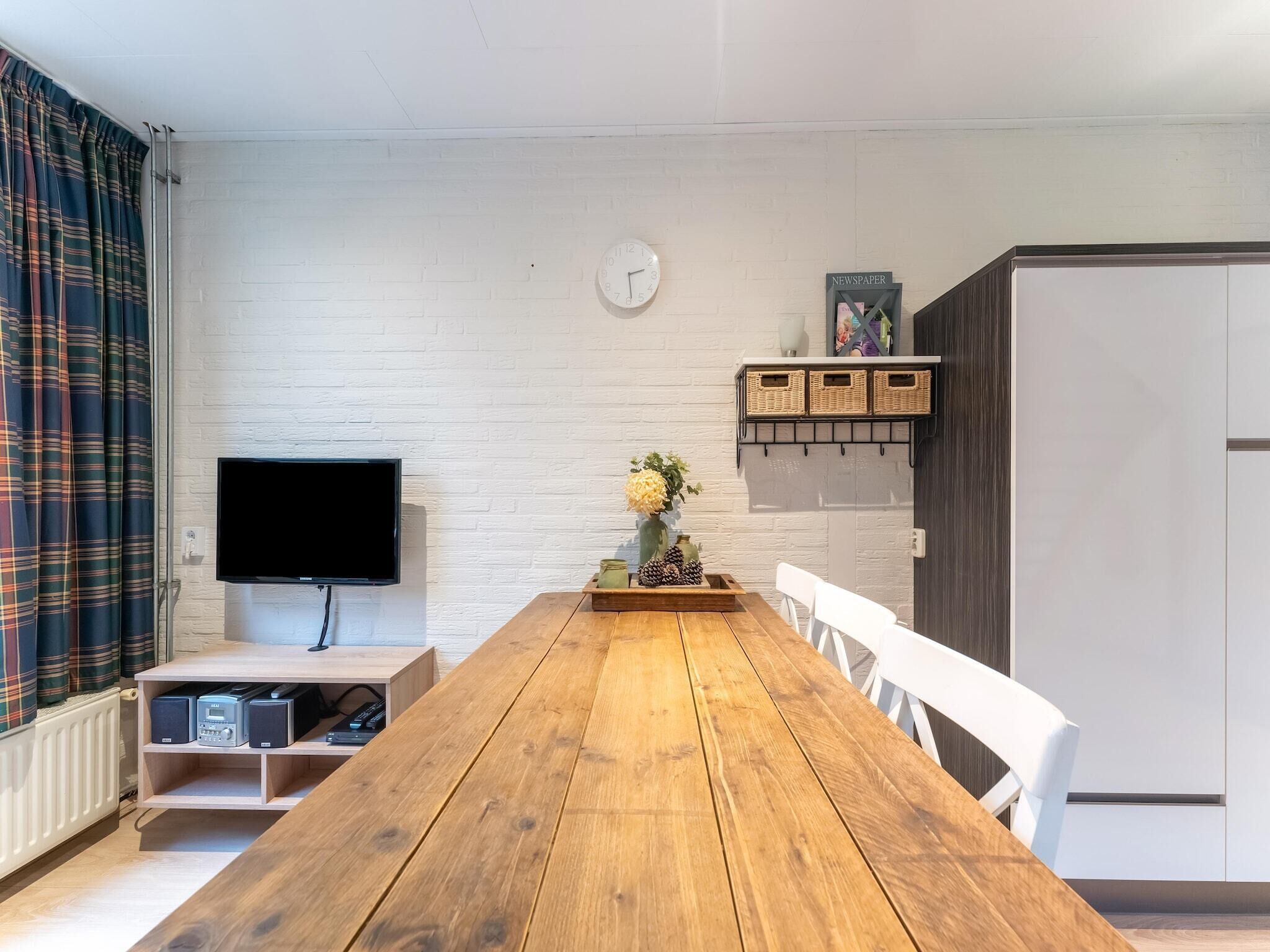 Een moderne woonkamer met een houten eettafel, witte stoelen, een flatscreen televisie, een klok en een plank met rieten manden.