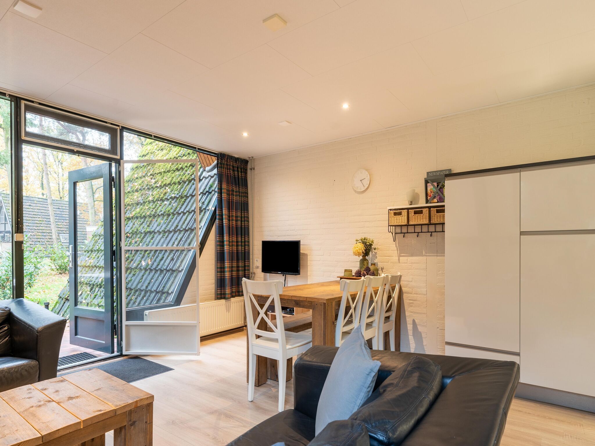 A modern living room with a sofa, a wooden table, and a dining area with chairs. There is a TV on a stand, a window with curtains, and a kitchen area with a refrigerator and a shelf with baskets.