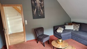 Un salon avec un canapé gris, un fauteuil gris, une table basse ronde ornée d’un objet en laiton, et une œuvre d’art représentant un éléphant, encadrée, accrochée au mur.