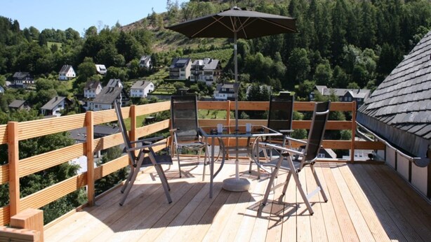 Une terrasse en bois, dotée de mobilier d’extérieur et d’un parasol, qui offre une vue sur une colline parsemée de maisons.
