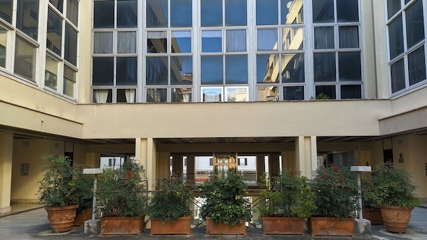 Edifício com várias janelas e área de entrada coberta, decorada com plantas em vasos.