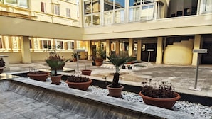 Um pátio com plantas em vasos dispostas ao longo de um caminho central, ladeado por um edifício com vários andares e varandas.