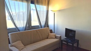 Uma sala de estar com um sofá bege, uma mesinha com televisão e controle remoto, e um abajur.