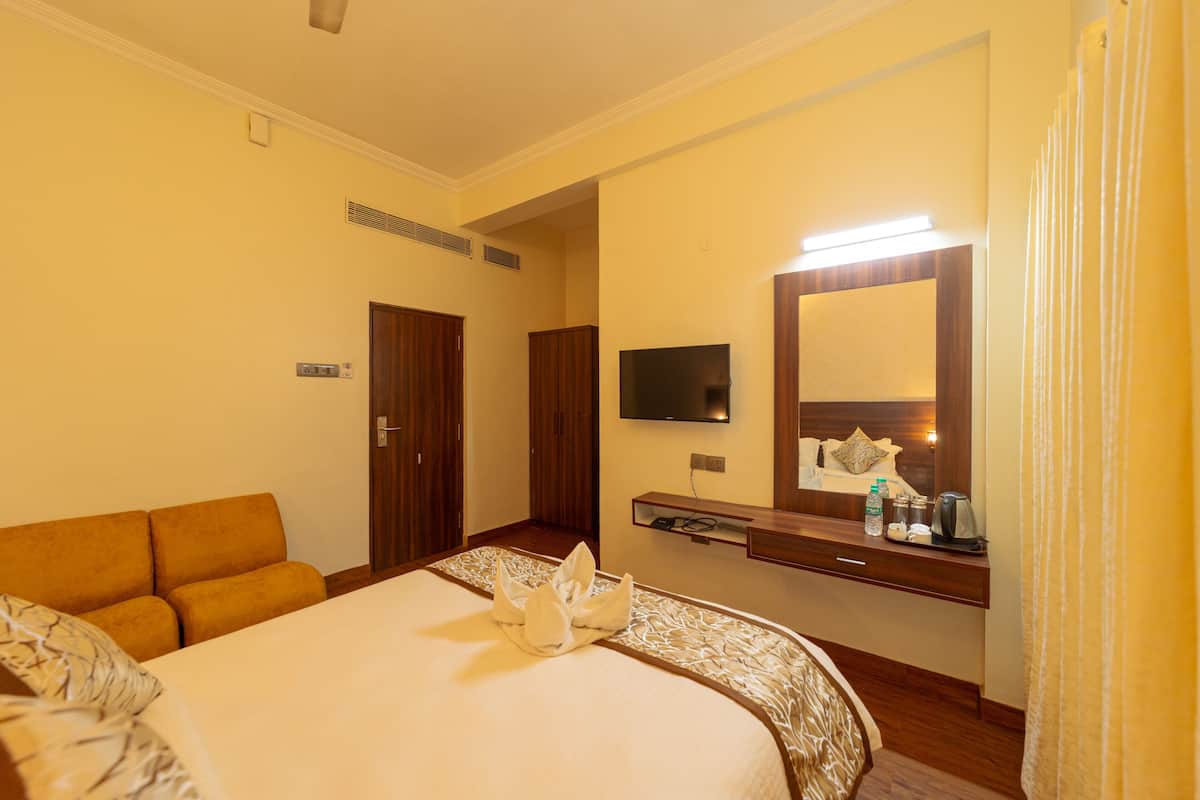 Una habitación de hotel con una cama, un sofá, un televisor, un espejo y una mesita de noche con una botella y una tetera.