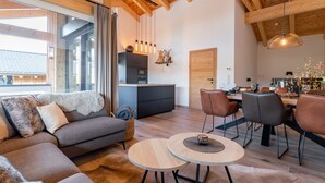 Un salon moderne avec un canapé, une table à manger et des chaises. Il y a un coin cuisine avec un réfrigérateur et une porte. La pièce est dotée de poutres en bois au plafond et de grandes fenêtres.