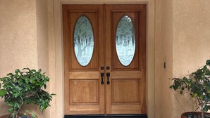 Una doble puerta de madera con paneles de vidrio, flanqueada por macetas con plantas, que da acceso a un vestíbulo con suelo de piedra.