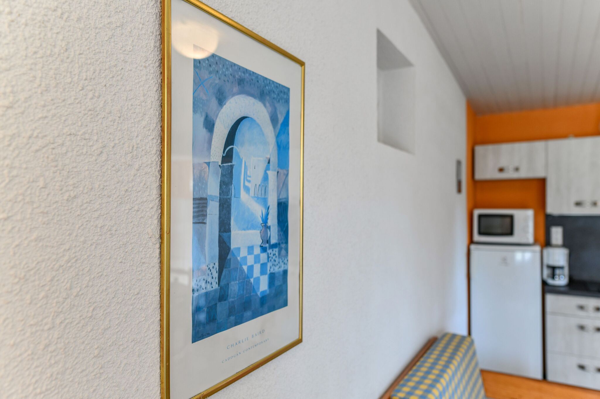 A framed artwork on a white textured wall, a kitchen area with white cabinets and appliances, and a patterned bench.