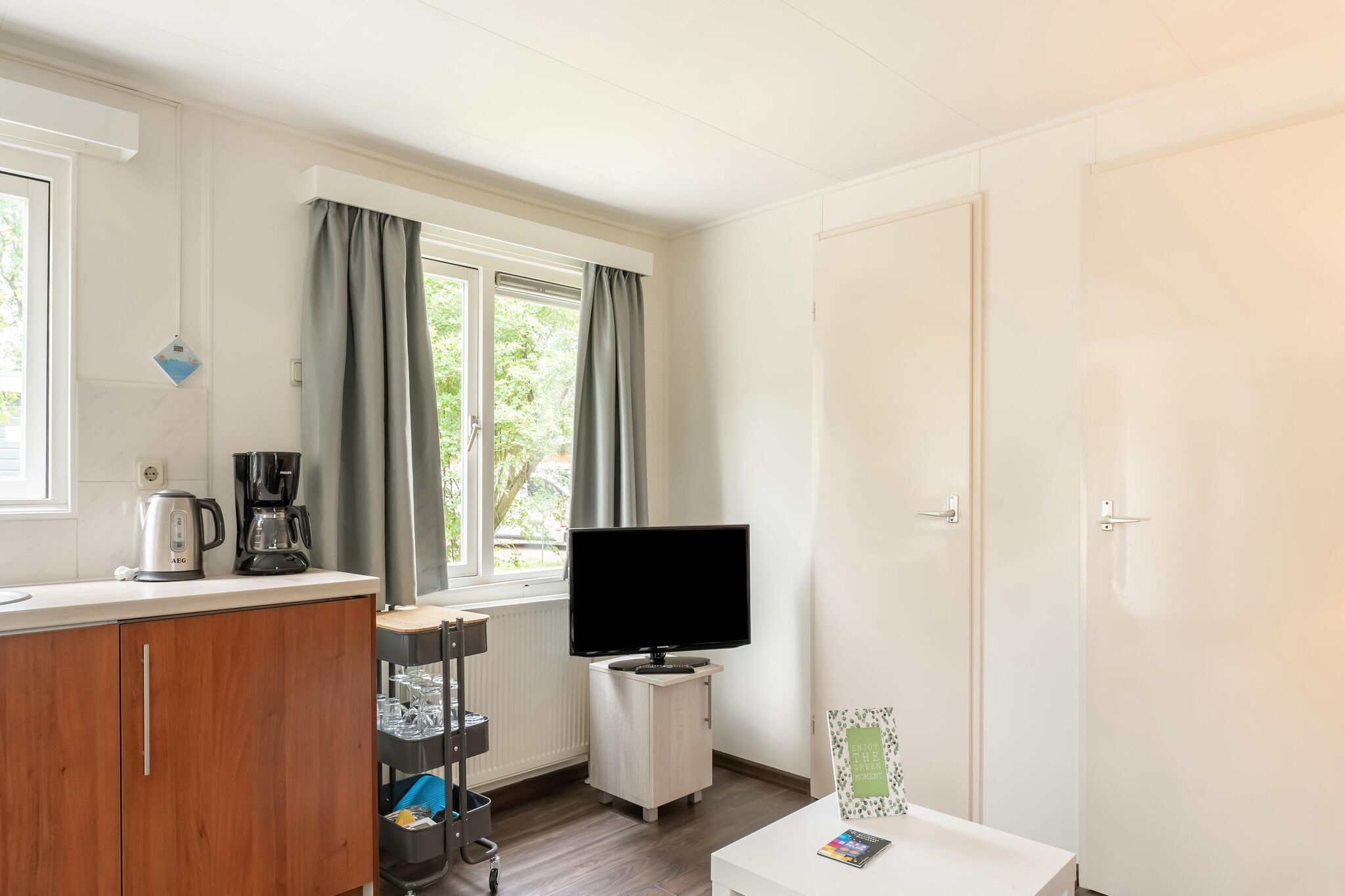 Una habitación con kitchenette, un televisor sobre un mueble, una ventana con cortinas y una puerta.