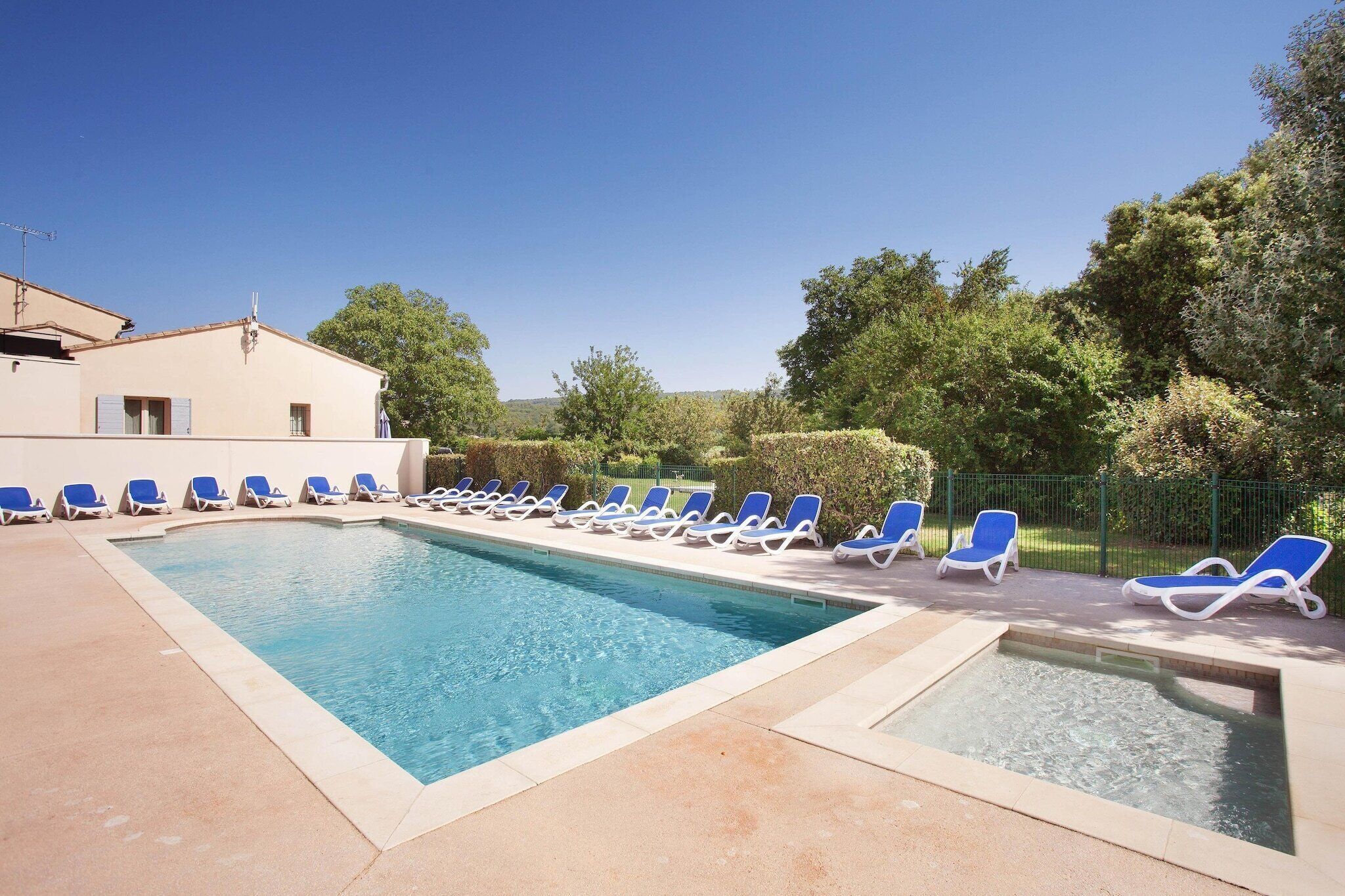 A rectangular swimming pool with blue water, surrounded by lounge chairs and a building with a light-colored facade.