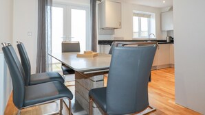 A modern kitchen with a dining area, featuring a table with chairs, a window with curtains, and a view of the outside. - 6B The Mews (Padstow)