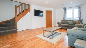 A living room with a wooden staircase, a grey sofa, a coffee table, and a TV mounted on the wall. - 6B The Mews (Padstow)