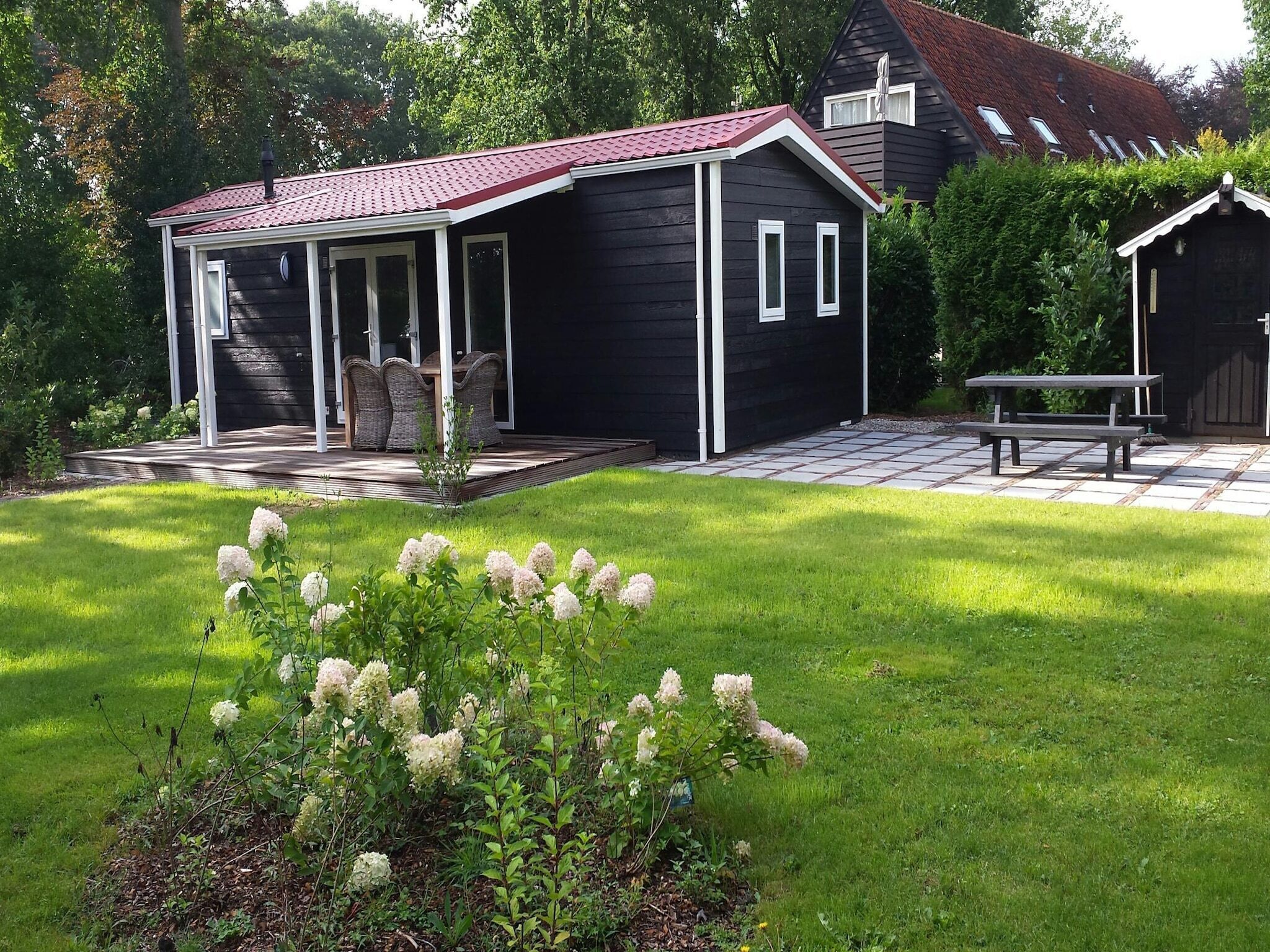 Une maison noire avec un toit rouge, une terrasse couverte et un petit jardin agrémenté de fleurs blanches.