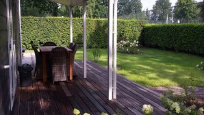 A covered patio with a wooden deck, a wicker table and chairs, and a garden with a hedge and flowers. - Peaceful Forest Chalet (Elst)