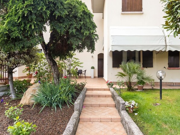A courtyard with a paved pathway, potted plants, a tree, and a building with a white facade and windows.