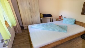 Una cama bien hecha con un cojín estampado en azul, un armario de madera, una mesita con una lámpara y una maleta en el suelo.