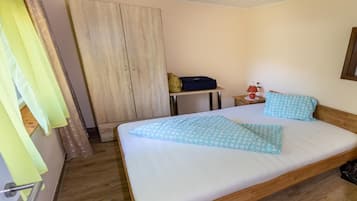 Een netjes opgemaakt bed met een blauw gestreept kussen, een houten garderobe, een klein tafeltje met een lamp en een koffer op de vloer.