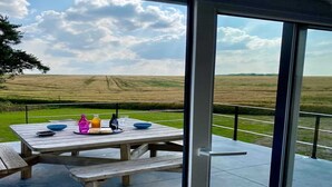 A wooden picnic table with a glass top, set on a patio with a view of a grassy field and distant hills. - Peaceful Garden Home in Modave (Modave)