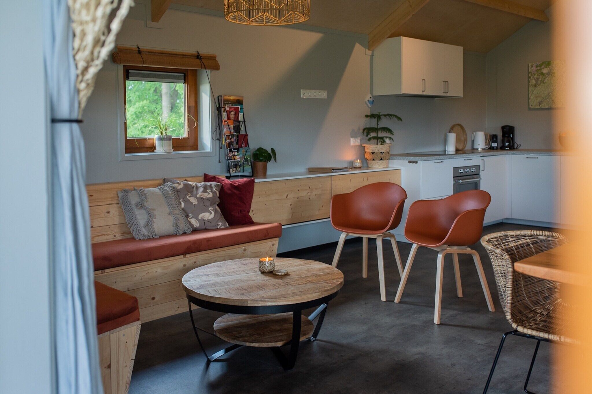 Une pièce chaleureuse avec un banc en bois, deux chaises, une table ronde avec une bougie, et un coin cuisine équipé de placards et d’une machine à café.