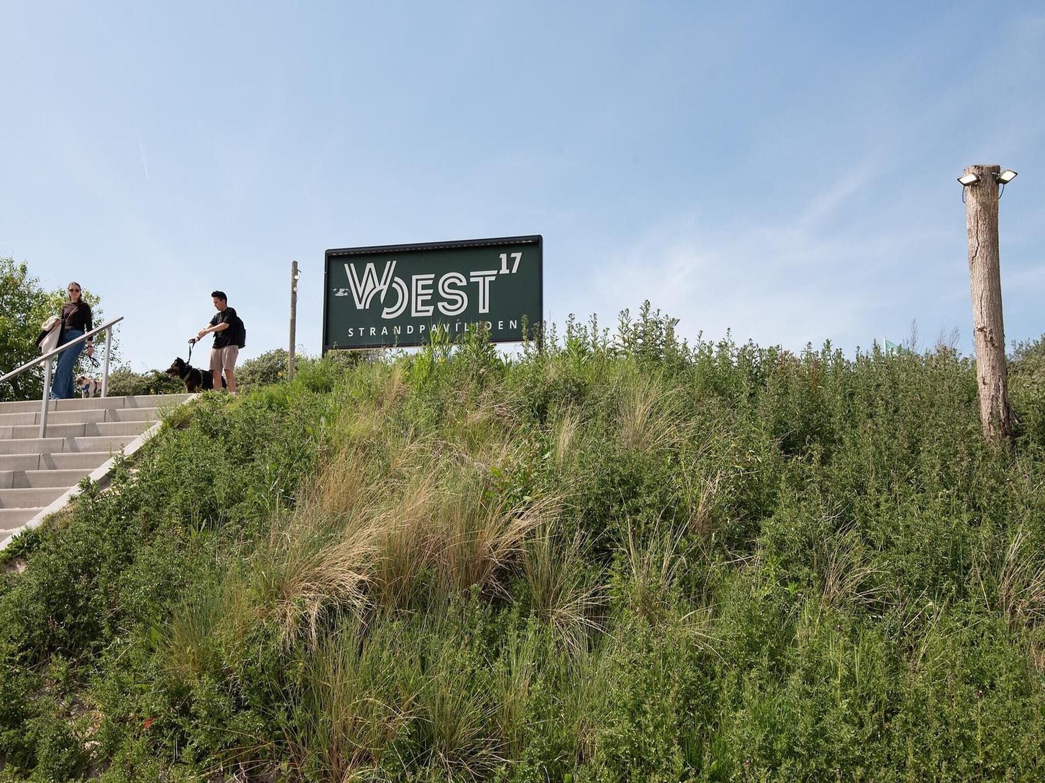 En skylt med texten ”WEST 17 STRANDPAVILIOEN”, med en trappa som leder upp till den, omgiven av vildvuxen vegetation.