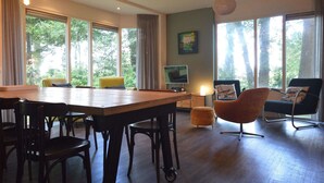 A spacious living area with a large wooden table, multiple chairs, and a television. Large windows provide ample natural light and views of trees. - Holiday Home in Lemele near Nature Reserve (Overijssel)