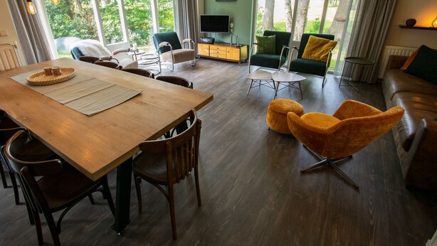A modern living room with a wooden dining table, comfortable seating, and large windows offering a view of greenery. - Holiday Home in Lemele near Nature Reserve (Overijssel)