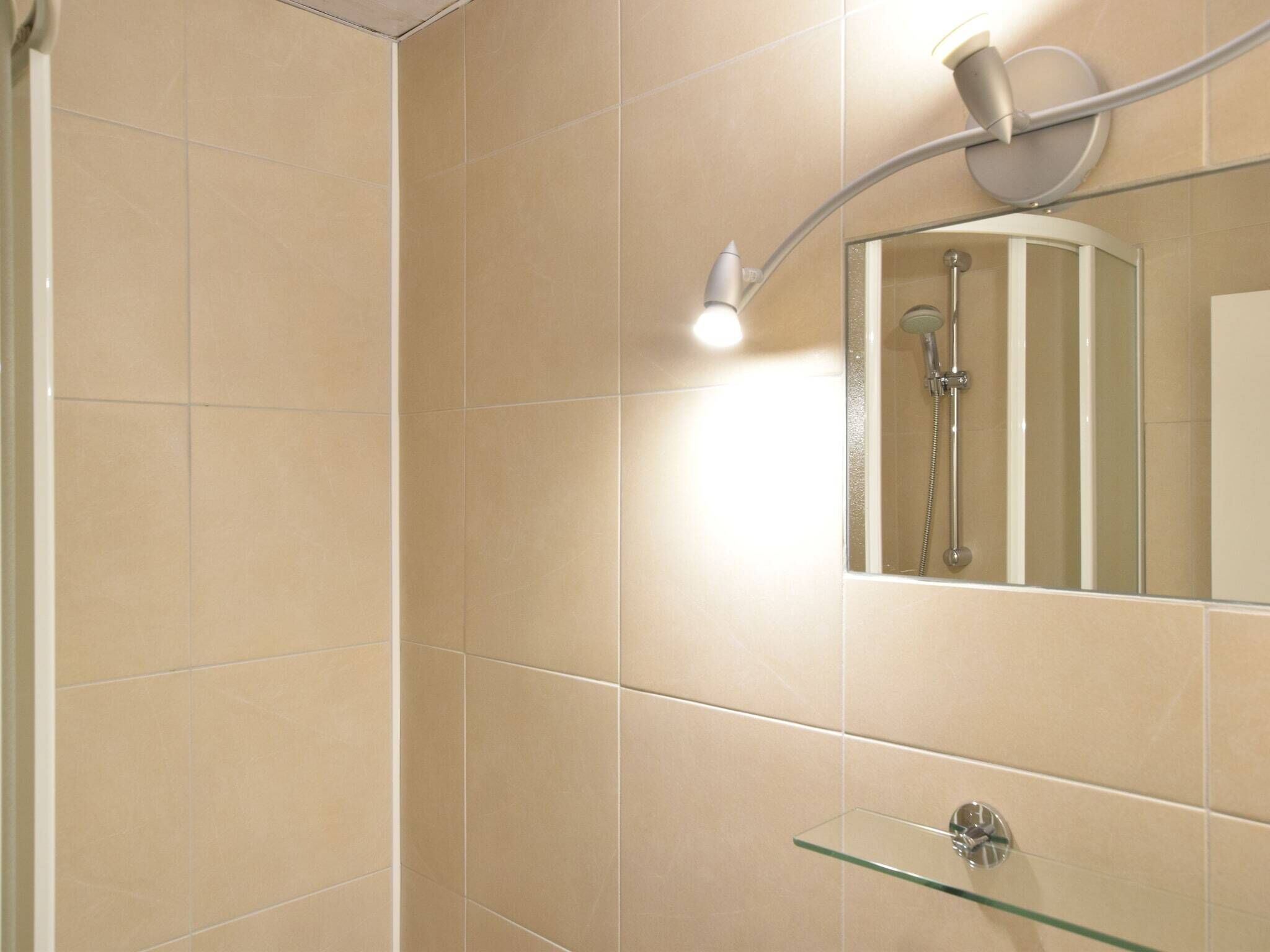 A modern bathroom with beige tiles, a glass shower enclosure, a mounted mirror, and a glass shelf.