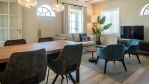 A modern living room with a dining area, featuring a large wooden table, comfortable seating, a television, and large windows. - Seals Cozy Farm Stay (De Cocksdorp Texel)