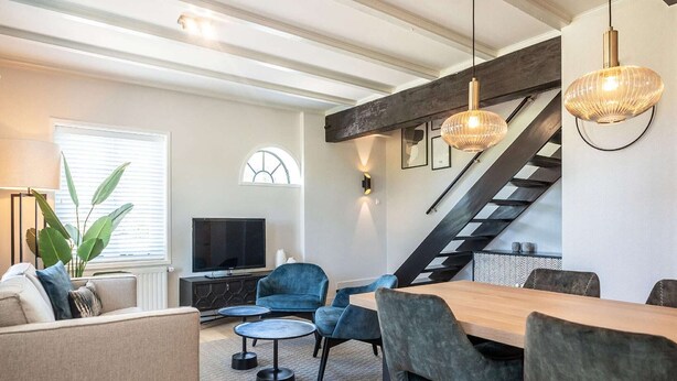A modern living room with a sofa, a TV, a dining table with chairs, a staircase, and a large window with blinds. - Seals Cozy Farm Stay (De Cocksdorp Texel)