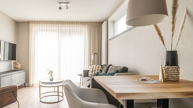 A modern living room with a sofa, a dining table, and chairs. There is a television on the wall, a window with curtains, and a vase with dried plants on the table.