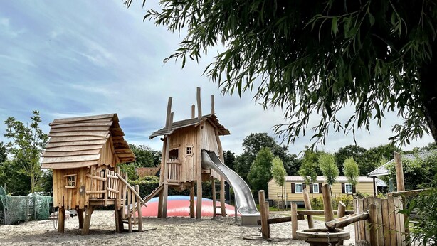 Een houten speelconstructie met een glijbaan en een klein huisje, omringd door zand en bomen.