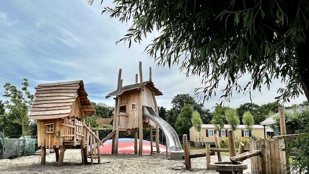 Een houten speelconstructie met een glijbaan en een klein huisje, omringd door zand en bomen.
