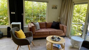 Een woonkamer met een bruine bank, een grijze fauteuil met een geel kussen, een ronde houten salontafel en een rieten voetenbankje.