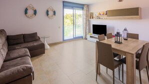 A modern living room with a sofa, a dining table with chairs, a TV, and a balcony with a view. - Belvilla by OYO Apartamento Augusta (Cambrils)