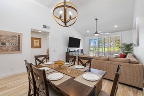 A dining area with a wooden table set for four, a ceiling fan, and a large window with a view of a green landscape. - Modern & Relaxing Condo | Stunning Fairway and Mountain Views | Ideal Vacation Retreat (Palm Desert)