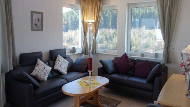 A living room with two black sofas, a wooden coffee table, and a lamp. There are windows with curtains, a framed picture on the wall, and a vase with flowers on a table. - Apartment in Willingen mit Balkon (Willingen)