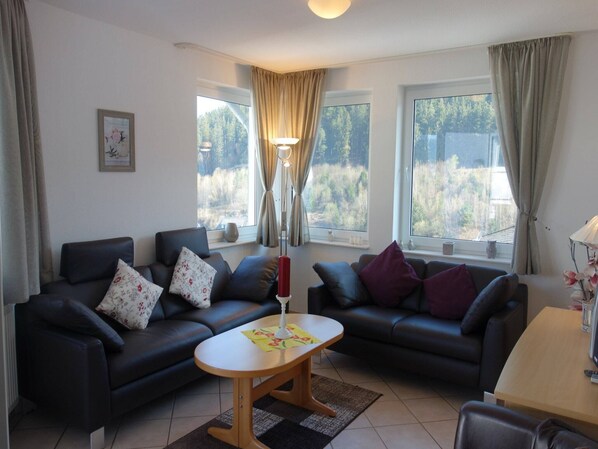 A living room with two black sofas, a wooden coffee table, and a lamp. There are windows with curtains, a framed picture on the wall, and a vase with flowers on a table. - Apartment in Willingen mit Balkon (Willingen)