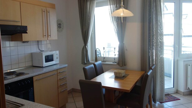 A compact kitchen with a microwave, stove, and wooden cabinets. There is a dining table with chairs and a window with curtains.