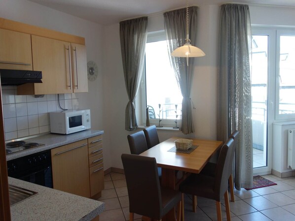 A compact kitchen with a microwave, stove, and wooden cabinets. There is a dining table with chairs and a window with curtains. - Apartment in Willingen mit Balkon (Willingen)