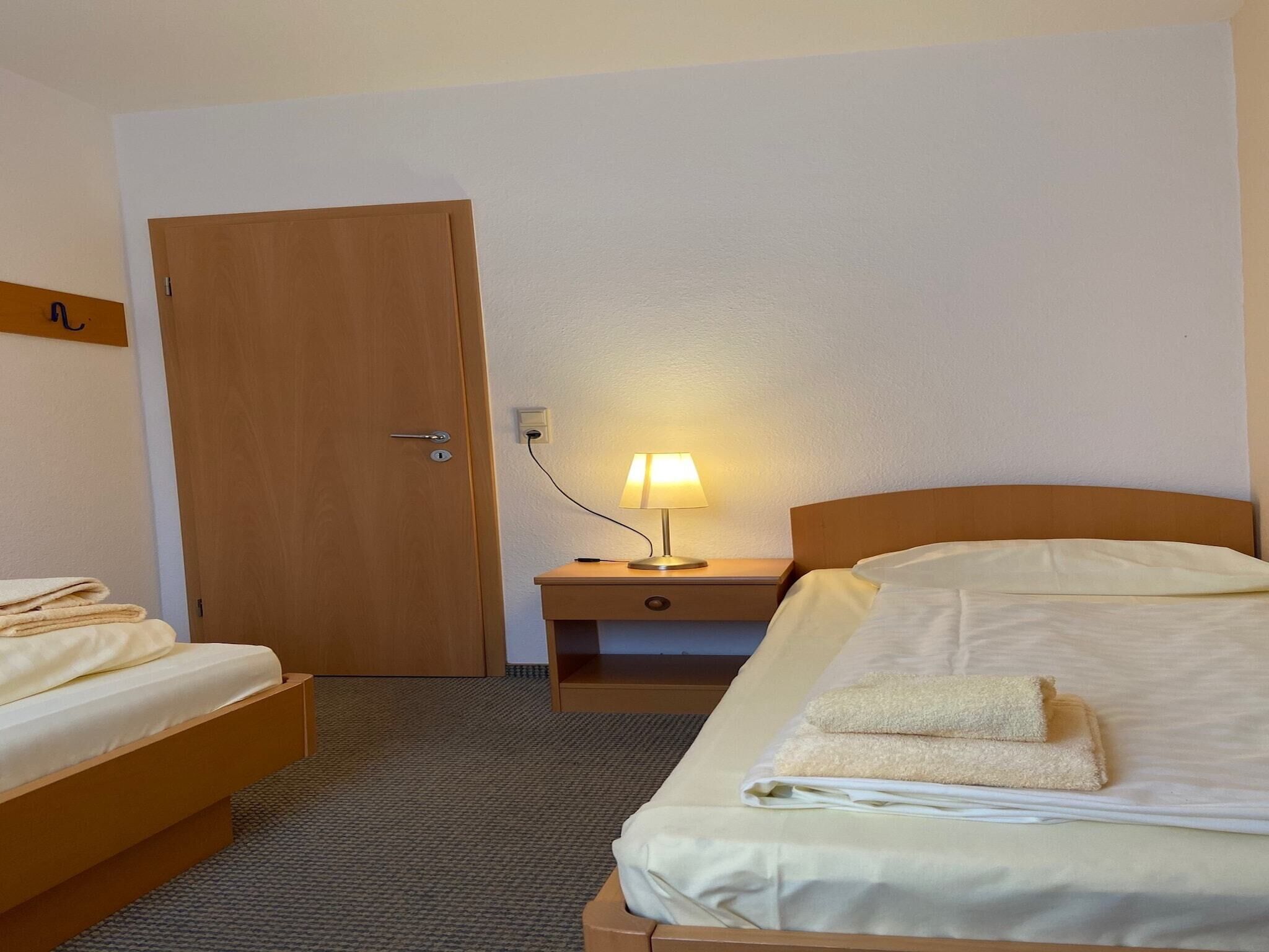 A hotel room with a bed, a nightstand with a lamp, a wooden door, and a small table with towels.
