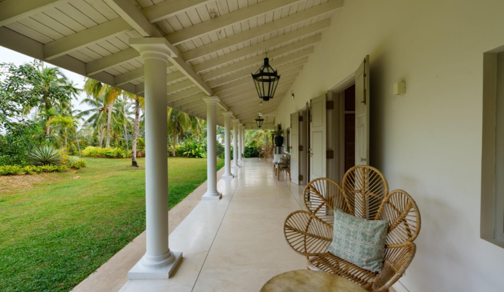 A covered porch with white columns, wicker chairs with cushions, and a view of a well-maintained garden with palm trees.