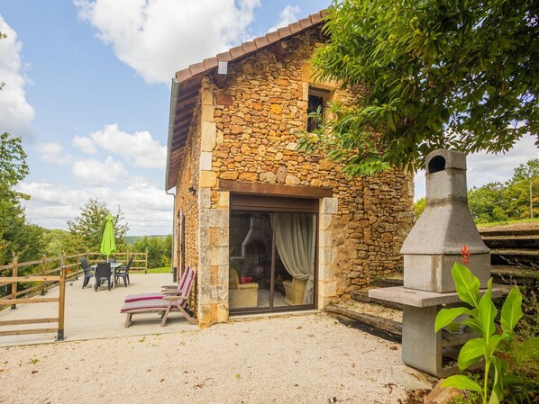 Un bâtiment en pierre doté d’une cheminée, d’un espace extérieur aménagé pour s’asseoir et d’une terrasse en gravier.