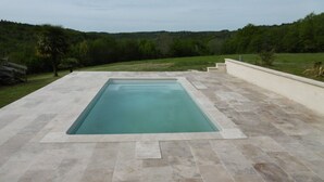 Une piscine rectangulaire bordée d’une terrasse en pierre, entourée d’une pelouse et d’arbres.