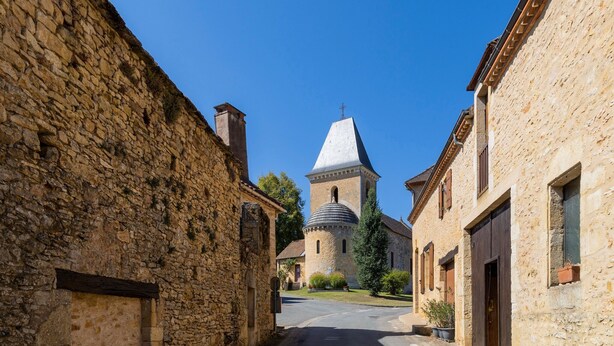 Une rue étroite bordée d’immeubles en pierre, une église dotée d’un clocher et un ciel d’un bleu limpide.