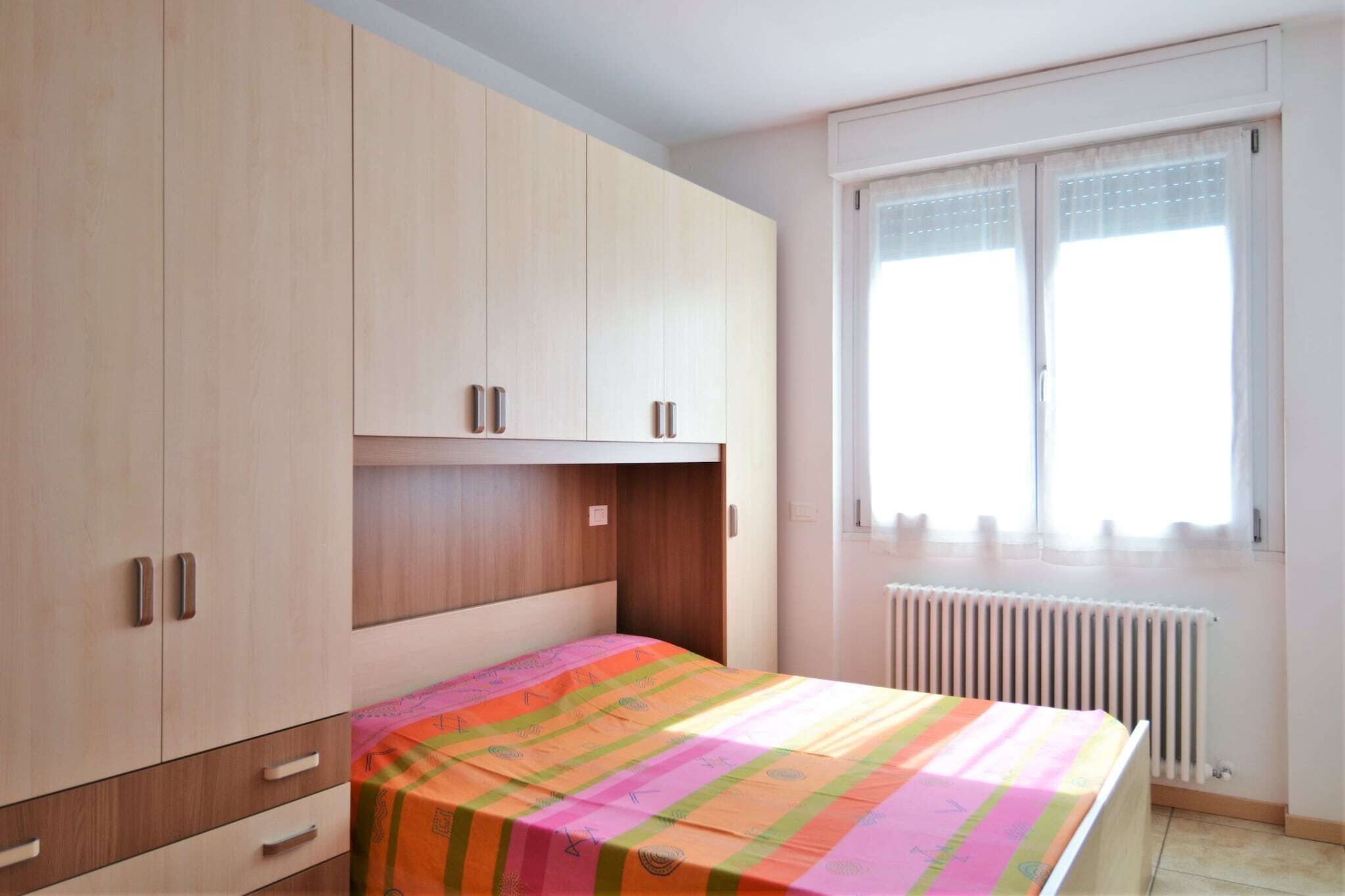 A bedroom with a bed, a window with curtains, a radiator, and wooden cabinets.