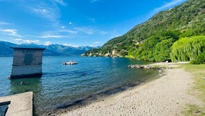 A lake with a clear view of a stone structure, a sandy beach, and a forested hillside. - Apartment by Lake Como with Scenic Views (Dervio)